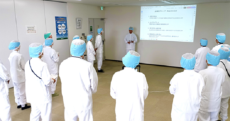 工場内で衛生服と帽子を着用した従業員が集まり、スクリーン資料を用いて食品安全に関するミーティングを行っている様子のイメージ